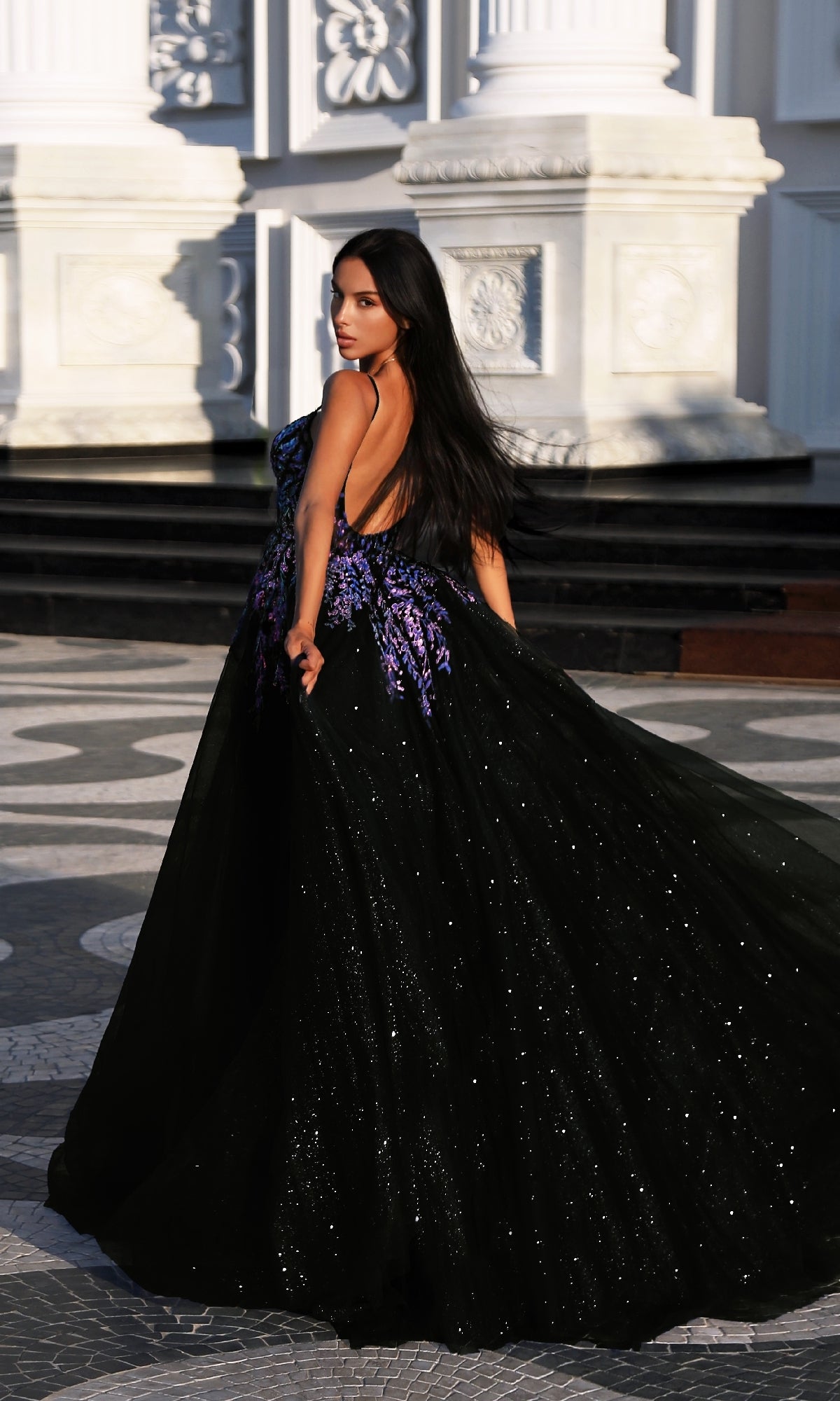 A woman with long dark hair models the Long Formal Dress: Tina Holly TE216B, a black and purple evening gown with floral appliques, standing outdoors on patterned stone in front of an ornate white building.